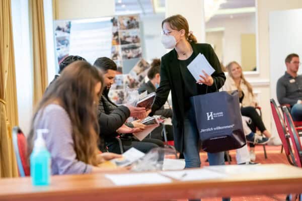 Hahne Holding Mitarbeiterin bei einer Veranstaltung, begrüßt Gäste mit Info-Material, Maskenpflicht, professionelle Unternehmenskommunikation.