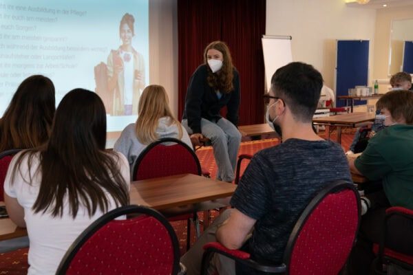 Verschiedene Schüler in einem Seminarraum beim Vortrag, alle tragen Masken, modernes Lernumfeld mit interaktiver Präsentation.