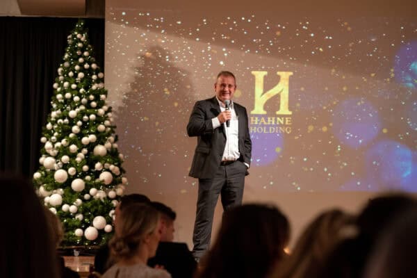 Moderner Geschäftsmann spricht bei Hahne Holding Event mit Weihnachtsbaum und Firmenlogo im Hintergrund.