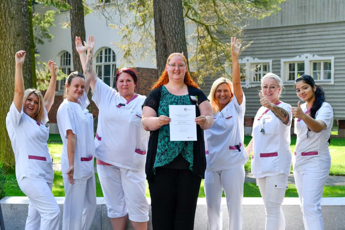 Gelbe Frauen in Pflegeuniform mit Urkunde vor Haus und Bäumen, Hahne Holding, Team, Erfolg, Pflege, Ausbildung, Mitarbeiter.