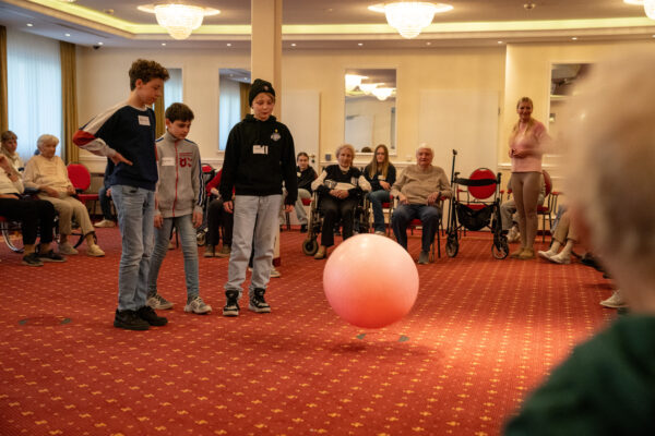 Mehrere Jugendliche und Senioren sitzen in einem großen Raum im Kreis, ein großer Gymnastikball liegt in der Mitte.