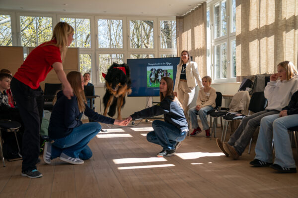 Hund springt durch die Arme zweier kniender Personen in einem sonnendurchfluteten Seminarraum, umgeben von einer Gruppe sitzender Menschen.